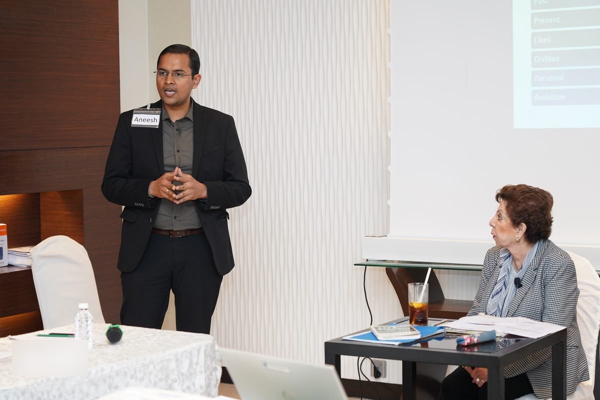 A participant speaks to the room while Sabira Merchant listens from her table